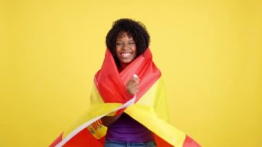 Stüdyoda sarı arka planda İspanyol bayrağı taşıyan afro saçlı bir göçmen kadın.