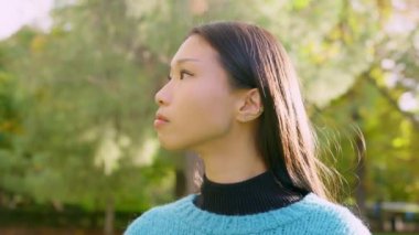 Ciddi bir Asyalı kadının parkta yan yan baktığı yavaş yörüngesel kamera görüntüsü.