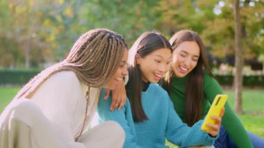 Üç çok kültürlü arkadaşın parkta cep telefonuna bakarken gülüşlerinin yavaş çekim videosu.