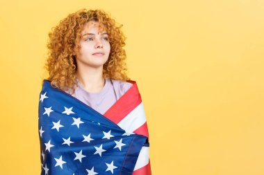 Stüdyo görüntüsü sarı arkaplan ve dikkat dağıtıcı kıvırcık saçlı kadın Kuzey Amerika ulusal bayrağıyla sarılı.