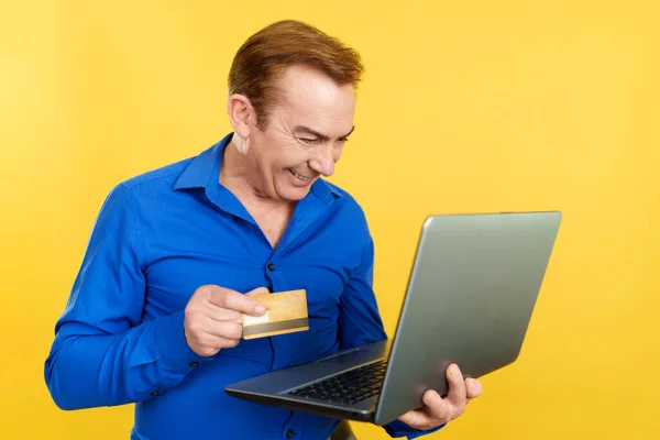 Mature man using a portable computer and card to shopping online in studio with yellow background