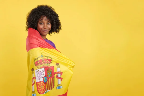 Migrant woman with afro hair wrapped with a Spanish flag in studio with yellow background