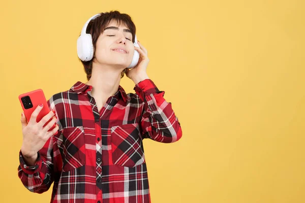 Relaxed androgynous person listening to music with mobile and headphones in studio with yellow background
