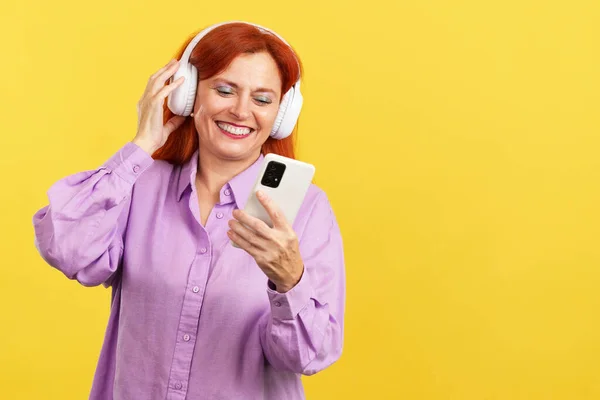 Mature woman with dye red hair listening to music using a mobile phone in studio with yellow background