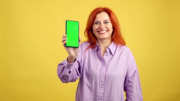 快乐红头发的成熟女人在有黄色背景的工作室里拿着带有彩色屏幕的手机 — 图库视频影像