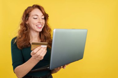 Kızıl saçlı kadın internetten laptop ve kredi kartıyla alışveriş yapıyor.