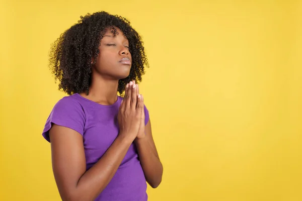 Afro saçlı bir kadın dua ediyor. Gözleri kapalı, elleri katlı, sarı arka planda.