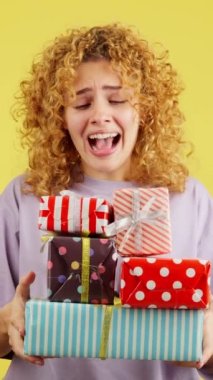 Studio video with yellow background of an excited young woman with curly hair and casual clothes receiving many gifts