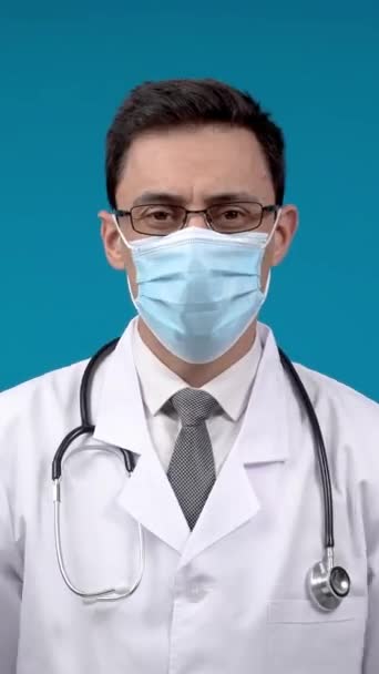 戴防护面罩的心满意足的医生交叉着胳膊 用听诊器在蓝色背景的脖子上看着相机 — 图库视频影像