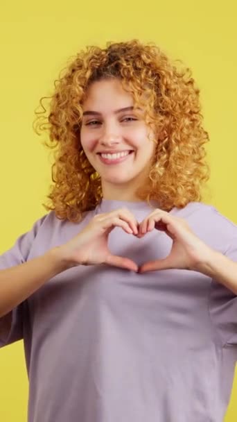 黄色の背景を持つスタジオビデオ幸せな若いです女性とともに巻き毛指の形で心を表す — ストック動画