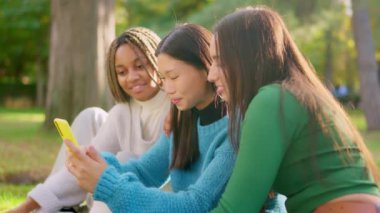 Cep telefonuna bakan ve parkın çimlerinde oturan çok ırklı arkadaşların yavaş çekim videosu.