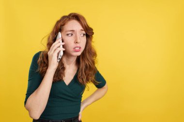 Stüdyoda sarı arka planda cep telefonuyla konuşan endişeli kızıl saçlı kadın.