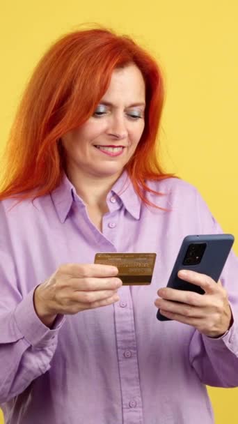 快乐成熟的红头发女人用手机和卡片在有黄色背景的工作室网上购物 — 图库视频影像
