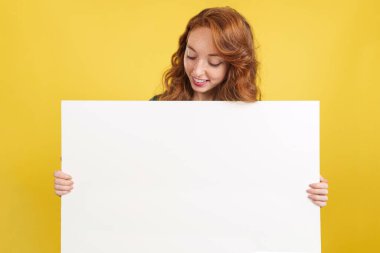 Kızıl saçlı kadın sarı arka planlı stüdyoda tuttuğu panele bakıyor.