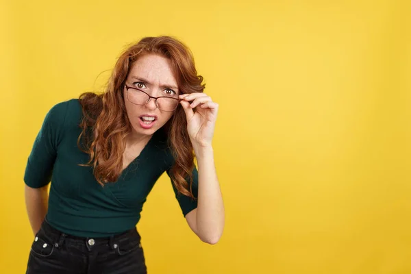 Redheaded woman with glasses staring at the camera in disbelief in studio with yellow background
