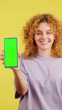 Studio video with yellow background of a smiley woman looking at camera while holding a mobile with the chroma screen