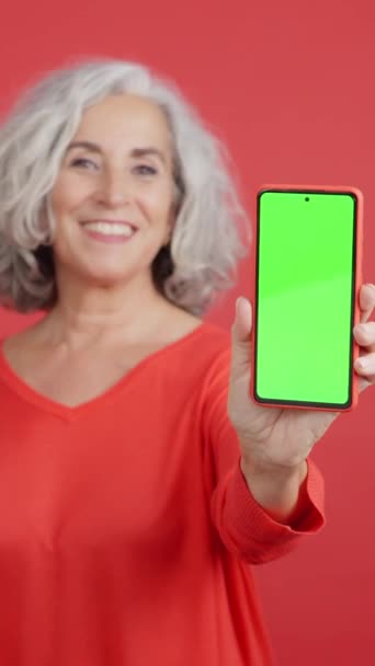 一个成熟的微笑女人拿着的带有红色背景的手机彩屏的工作室视频 — 图库视频影像