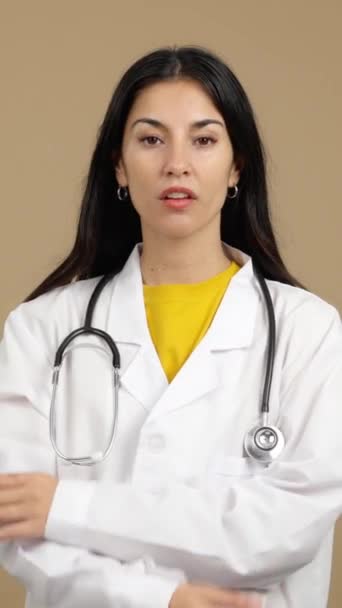 白人女性医師笑顔でカメラに腕を組んでスタジオで交差茶色の背景 — ストック動画