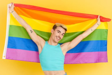 Gururlu gay adam sarı arka planlı stüdyoda bir lgbt gökkuşağı bayrağı dikiyor.