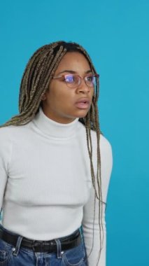 Video with blue background of an african woman with eyeglasses staring at camera in disbelief, side view