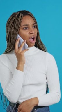 Video with blue background of an african woman with a surprised expression talking on the mobile phone