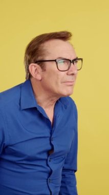 Olgun bir adam, sarı arka planlı stüdyoyu görmek için gözlüklerini çıkarıyor.