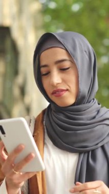 Mutlu Müslüman kadın yürürken gülümsüyor ve sokakta cep telefonu kullanıyor.