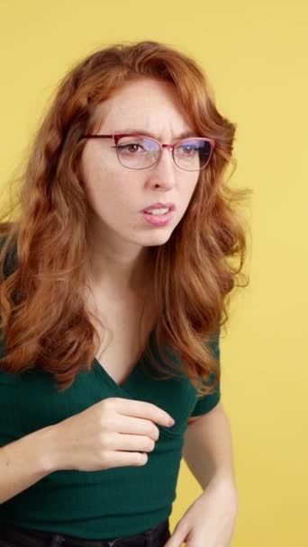 带着眼镜的红头发女人好奇地在有黄色背景的工作室里窥探 — 图库视频影像