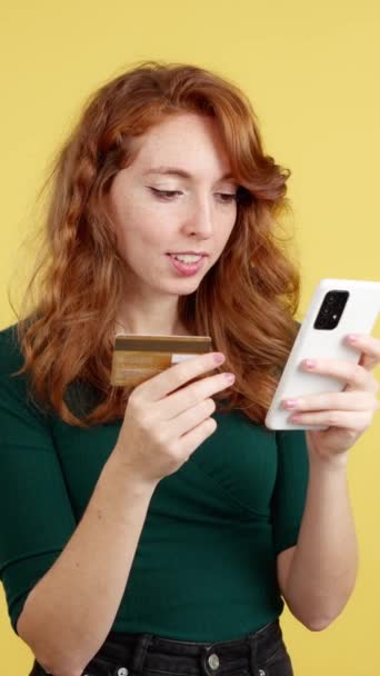 快乐的红头发女人带着手机和一张黄色背景的卡片在网上购物 — 图库视频影像