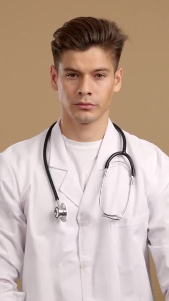 Przystojny Poważny Lekarz Patrzący Kamerę Stojącą Rękami Skrzyżowanymi Studiu Brązowym — Wideo stockowe