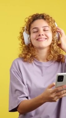Studio video with yellow background of a smiley woman with curly hair listening to music with a mobile and headphones