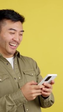 Happy chinese man smiling while using the mobile in studio with yellow background