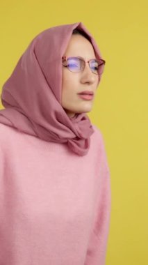 Müslüman kadın, sarı arka planlı stüdyoya bakarken gözlüklerini çıkarıyor.