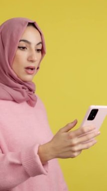 Cep telefonunun ekranına bakan şaşırmış Müslüman bir kadının sarı arka planlı stüdyo görüntüsü