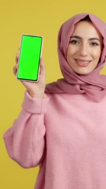 Stüdyoda sarı arka planda krom ekranlı cep telefonu tutan mutlu Müslüman kadın.