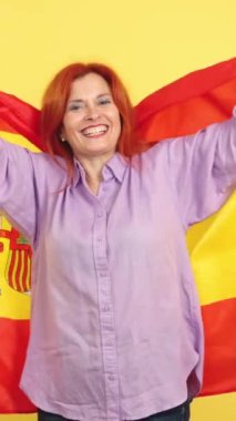 Stüdyoda sarı arka planda İspanyol ulusal bayrağıyla sarılan mutlu kızıl saçlı olgun kadın.