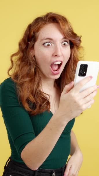 Удивлённая Рыжая Женщина Смотрящая Экран Мобильного Телефона Студии Жёлтым Фоном — стоковое видео