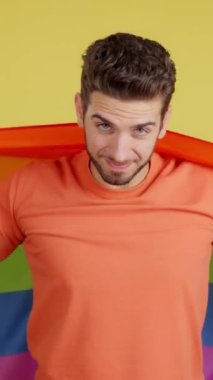 Stüdyoda sarı arka planda lgbt bayrağıyla sarılı mutlu beyaz adam.