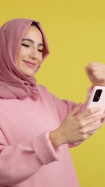 Excited muslim woman celebrating raising the fist while using a mobile in studio with yellow background