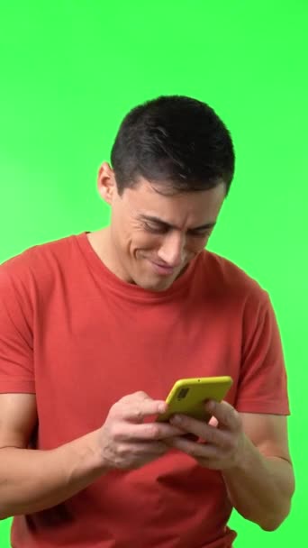 Tシャツに濃い髪の若い男性が楽しそうに笑いながらスマホを使いながら緑を背景に良いメッセージを受け取る内容 — ストック動画