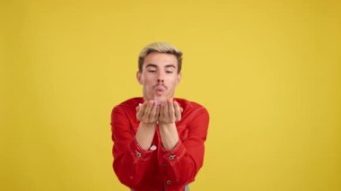 Stüdyoda sarı arka planda konfeti üfleyen mutlu gay adam.