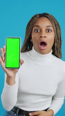 Video with blue background of an african woman with a surprised look on her face showing a mobile phone with chroma screen