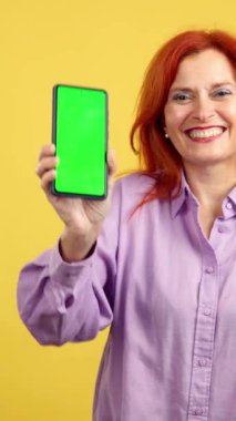 Stüdyoda sarı arka planda krom ekranlı cep telefonu tutan mutlu kızıl saçlı olgun kadın.
