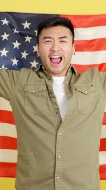 Happy chinese man raising a USA national flag while looking at camera in studio with yellow background