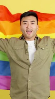 Happy chinese man raising a lgbt rainbow flag in studio with yellow background