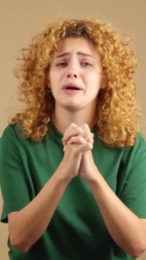 Studio video of woman beggong with curly hair and a worried face, praying with her hands clasped while looking into the camera