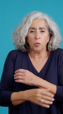 Studio video with blue background of an aged caucasian woman looking at camera while embracing herself expressing coldness