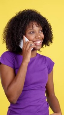Happy woman with afro hair smiling while talking to the mobile in studio with yellow background