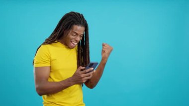 Stüdyoda mavi arka planda cep telefonunu kullanırken rastalı saçlı heyecanlı Latin adam kutlama yapıyor.