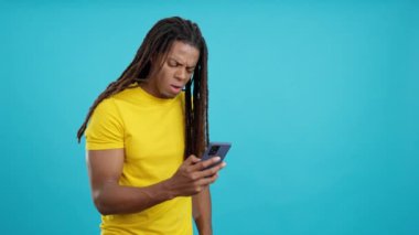 Stüdyoda mavi arka planda cep telefonu kullanan, rastalı, endişeli Latin adam.